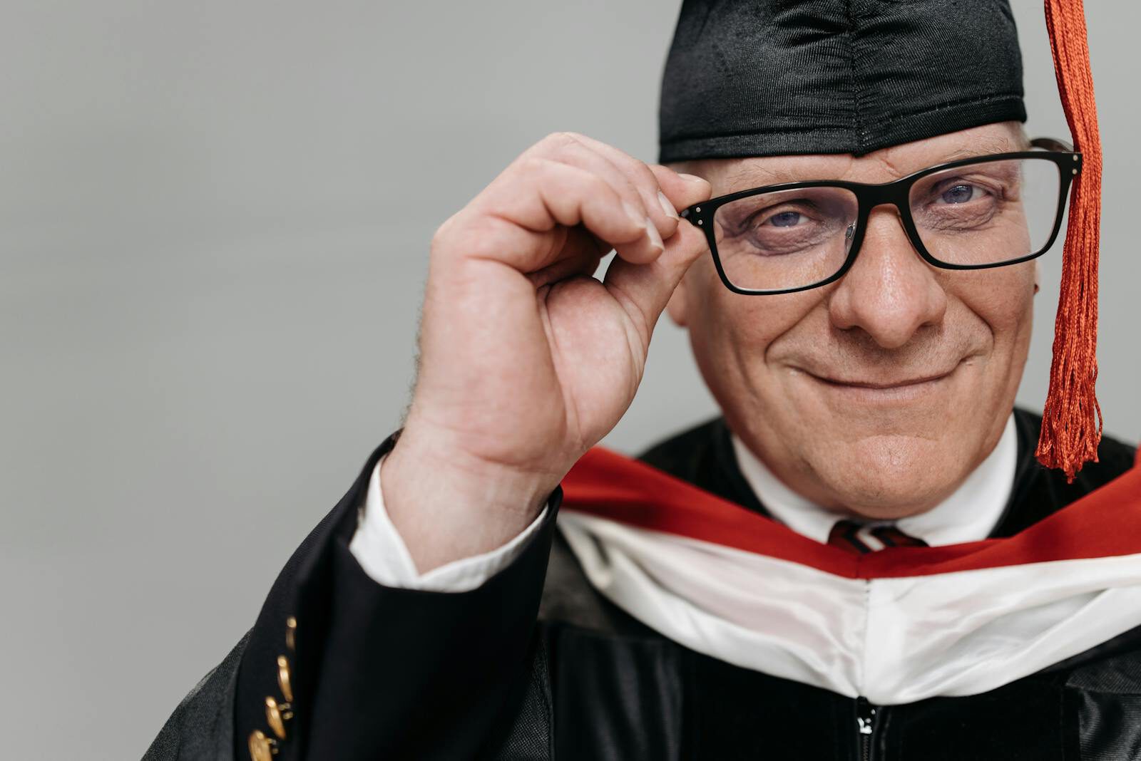 Elderly man in graduation attire happily posing with eyeglasses.